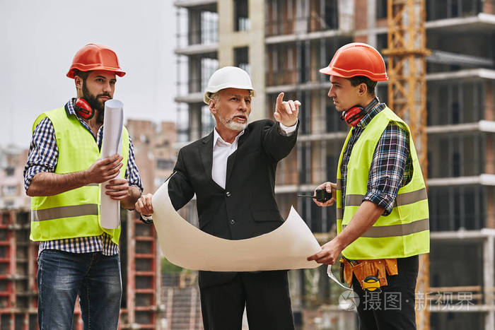 身着正装,头戴白盔的总工程师手持新项目草图,站在施工现场与两名年轻