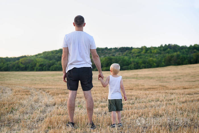 父亲和一个年幼的儿子站在割过的麦田上。后视图。日落时间