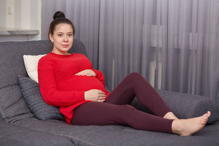 照片中的孕妇穿着红色毛衣和栗色长裤,手放在肚子上,模特看着镜头,坐