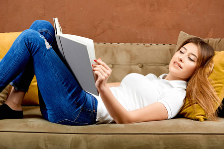 马球衫和蓝色牛仔裤的女人正靠着棕色的墙壁躺在棕色的沙发上看书照片