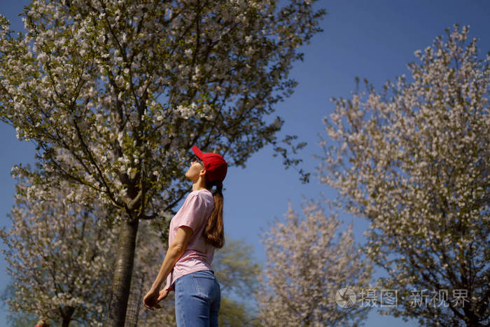 成功的商务女性喜欢在樱花盛开的公园里闲暇时光，穿着牛仔裤粉色t恤和时尚的红帽子