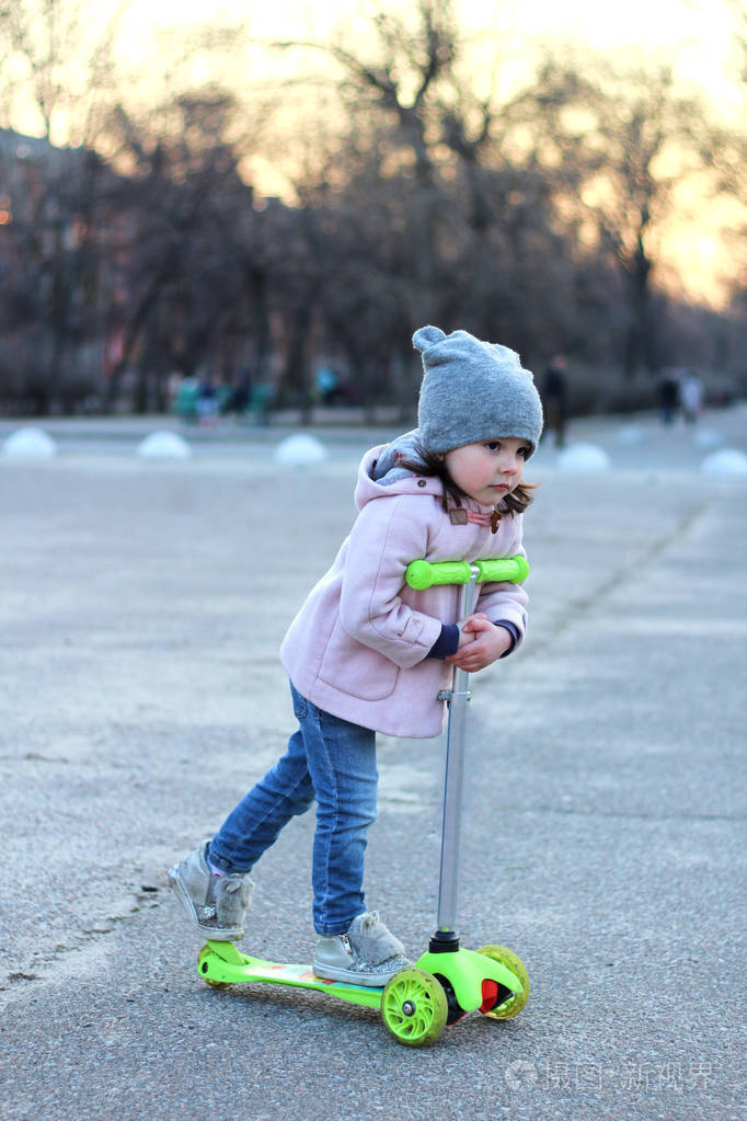 戴着帽子外套和牛仔裤的可爱女孩骑着三轮摩托车晚春城落日