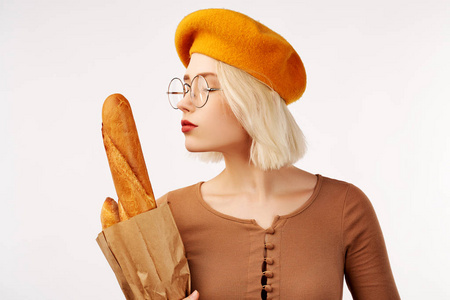 年轻快乐的金发女士戴着黄色贝雷帽,抱着包里的长棍面包,在工作室的