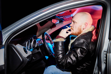 穿着皮夹克的男司机在黑暗中在车里抽烟.夜间拍照照片