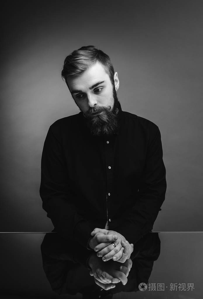 一张黑白照片一个留着胡子留着时髦发型的男子穿着黑色衬衫站在镜子旁