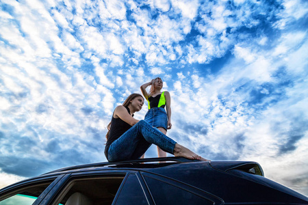 两个女孩坐在一辆黑色大轿车的车顶上,头顶上是白云摄影图