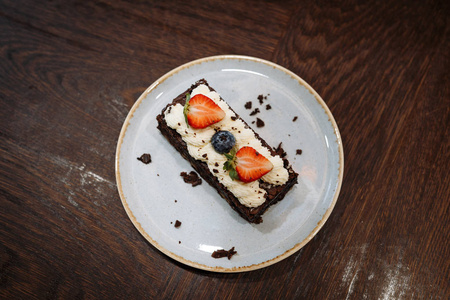 在深色木质背景的盘子里,用鲜奶油,草莓和蓝莓装饰的布朗尼蛋糕照片