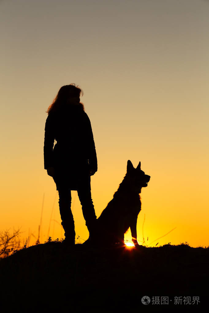 狗的剪影坐在附近的女人女孩自信地站在山上日落时在田野里的宠物概念