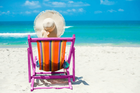 沙滩上,一个女孩坐在沙滩椅上,头戴彩色帕雷奥和草帽,背对着相机,放松