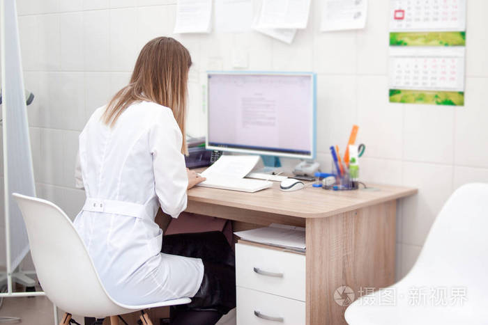 白种金发女医生在诊所医院的背面视图在电脑上工作.背景上有橱柜和其