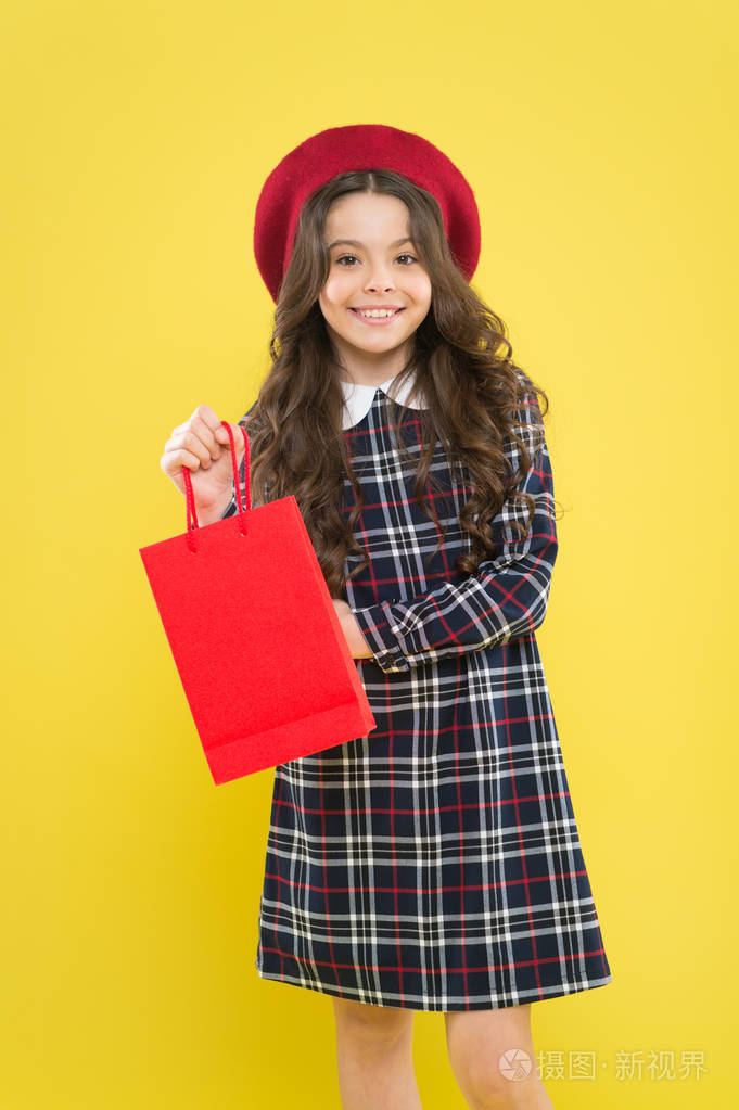 小女孩微笑着在黄色背景上买东西.快乐的小孩拿着红色纸袋买的东西.