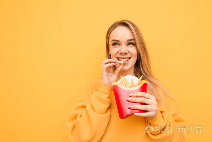 一个穿着橙色衣服微笑的女孩站在黄色背景上吃薯条,在一片空白处侧视.