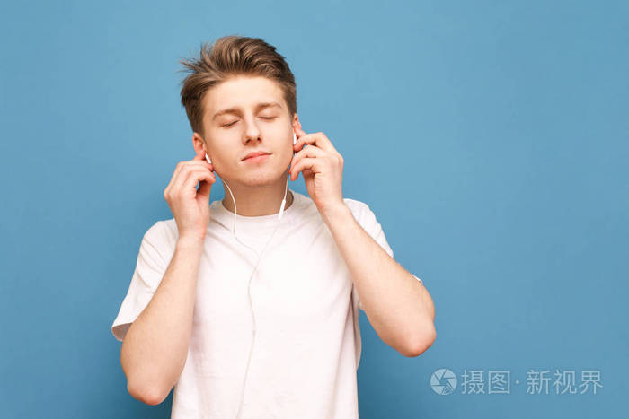 一个穿着白色t恤的漂亮男人闭着眼睛,戴着耳机听音乐,在蓝色背景下与