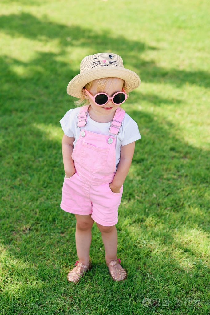 在阳光明媚的夏日，穿着短裙戴着帽子戴着粉色眼镜的可爱女孩站在绿草成荫的草坪上。