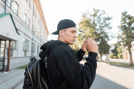 戴着棒球帽,戴着太阳镜,背着背包的年轻人正在一座老式建筑附近散步