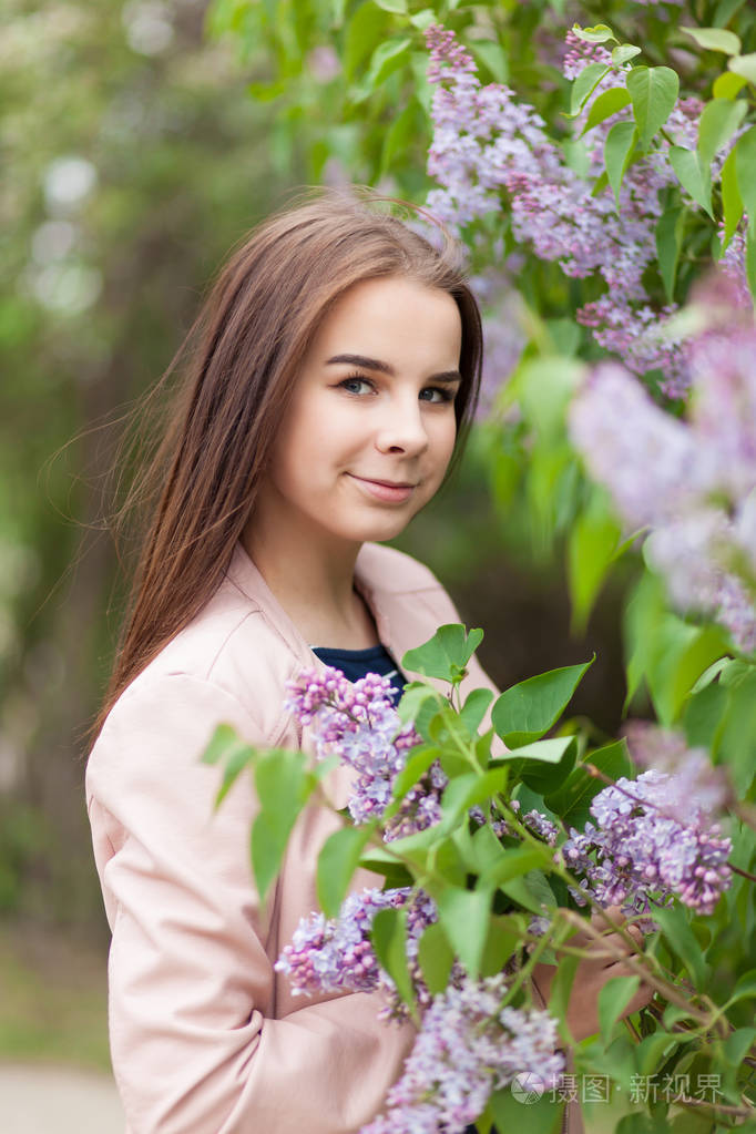 年轻漂亮的女孩站在盛开的丁香花丛旁