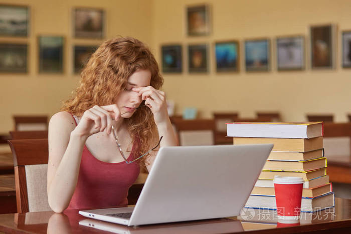 悲伤的女人她带着笔记本电脑坐在阅览室的桌子旁戴着眼镜手指放在鼻子