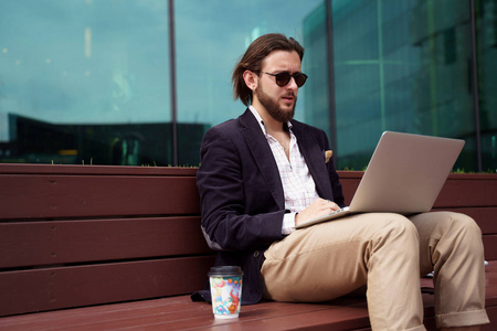 图为一位男性自由职业者拿着笔记本电脑坐在玻璃墙旁的木凳上照片