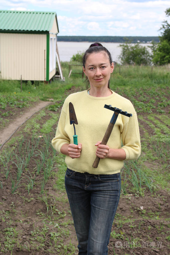 穿着黄色羊毛衫和牛仔裤的农业美女拿着铲子和花园耙子在田里园丁在