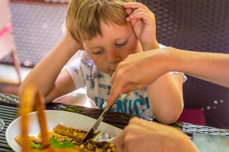 一个5-6岁的小男孩正在餐馆里吃土豆泥和肉排.妈妈切肉饼.照片