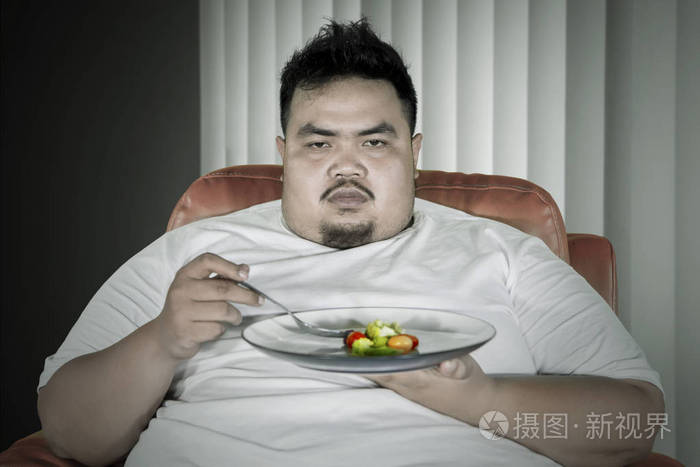闷闷不乐的肥胖男人在沙发上吃沙拉