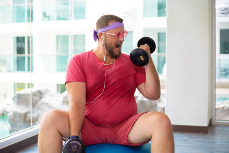 滑稽的胖男人戴着粉色眼镜,穿着粉色t恤,在健身房里用哑铃在健身球上