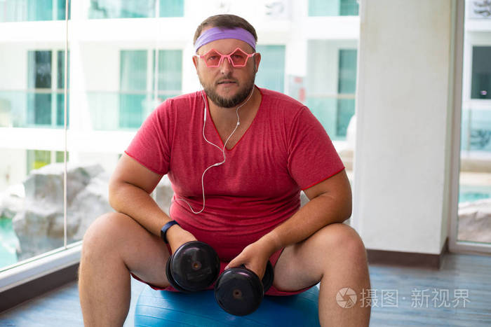 一个穿着粉色t恤和粉色眼镜的顽皮胖子正在健身房里用哑铃和健身球