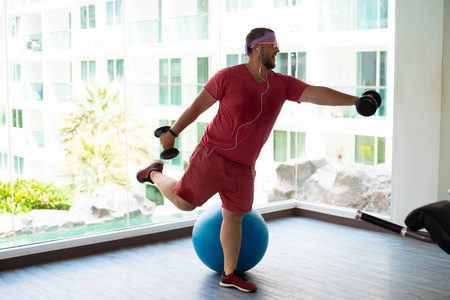 一个穿着粉色t恤和粉色眼镜的顽皮胖子正在健身房里用哑铃和健身球