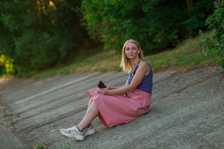 身穿紫色上衣,浅粉色短裙,穿着运动鞋的金发女孩坐在路面斜坡上,戴着