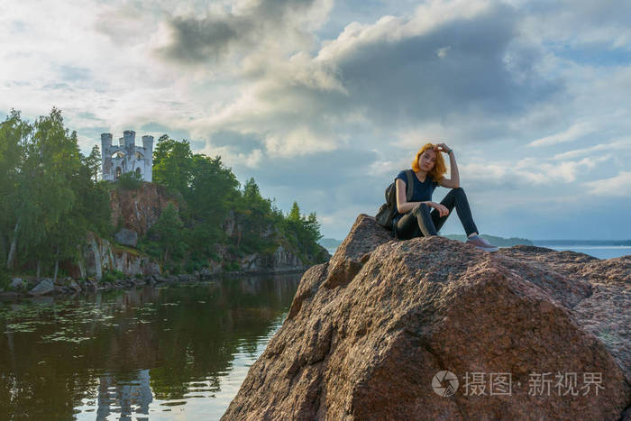 夏日傍晚，红发美女坐在海湾岸边的石头上。美丽的风景背景下的游客。蒙雷普斯公园，维堡，俄罗斯。旅游概念