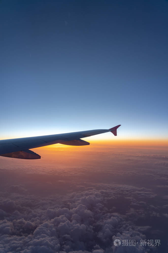 从飞机窗口观看飞机机翼轮廓在太阳升起时具有深蓝色的地平线和云背景