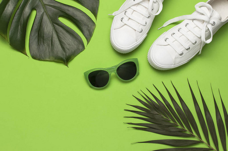 白色女式时尚运动鞋,时髦的太阳镜,绿色背景上的热带树叶.