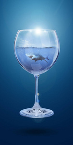 蓝色背景下,一杯葡萄酒中的鲨鱼拼贴画.酒精的危害.带海水的酒杯.