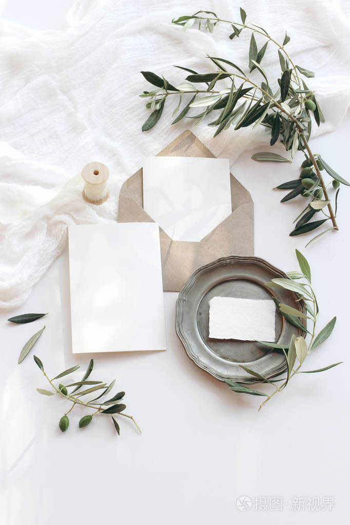 夏季婚礼文具模拟场景。空白贺卡信封复古银盘橄榄枝和丝带。白色背景，阳光下的棉制桌布。平视图。