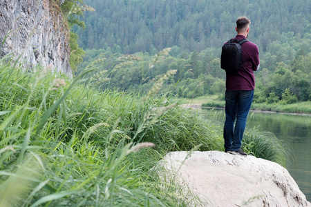 个穿着牛仔裤和衬衫背着背包的旅行者站在山河岸边的一块石头上向前看