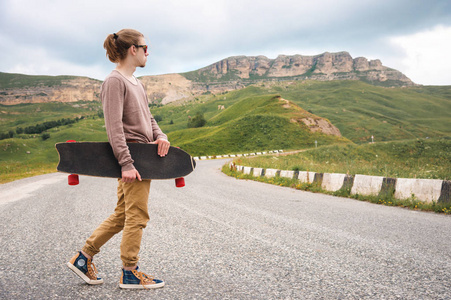 一个时髦的年轻人手里拿着溜冰鞋或长木板沿着蜿蜒的山路行走