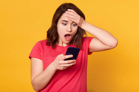 的女人,一只手放在额头上,拿着智能手机,惊讶地张大嘴巴,看着手机屏幕