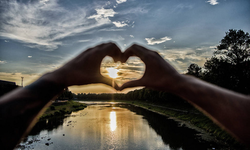 男性手呈心形手势,象征爱情和浪漫.河上日落前的心形照片