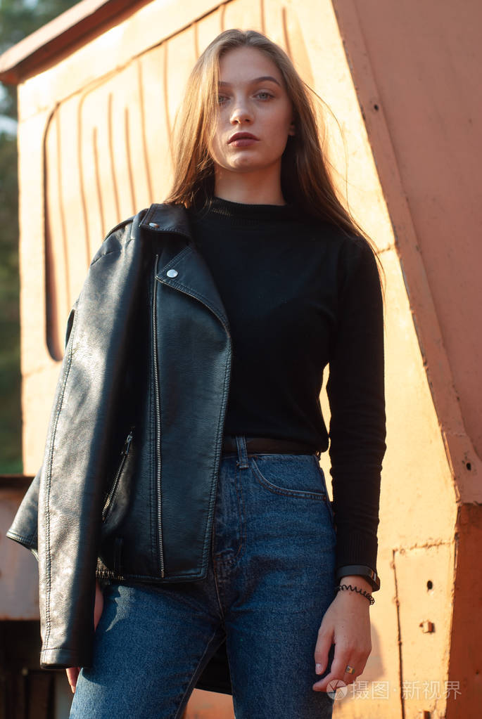穿着黑色皮夹克和蓝色牛仔裤的模特在户外摆姿势现代酷女孩