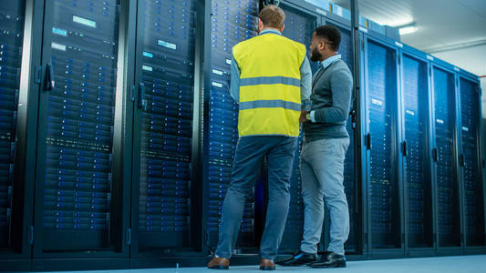 管理员穿着高能见度背心,手持笔记本电脑,站在服务器机架前,正在数据