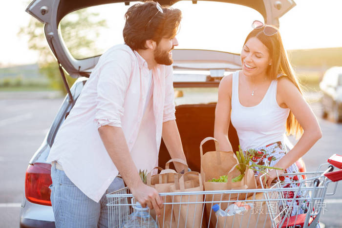 快乐的年轻夫妇从杂货店购物回来把装满食物的纸袋装进汽车后备箱