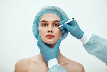 医疗帽等待着面部手术,而戴着蓝色手套的整形医生在脸上画着虚线照片