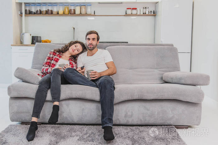 爱心的人手里拿着杯子妻子把头靠在肩上在看电视的时候坐在家里的沙发