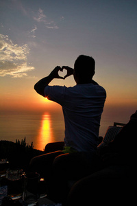 小男孩在海边欣赏日落,在阳光下观看并制作心形标志,阳光反射在海面上