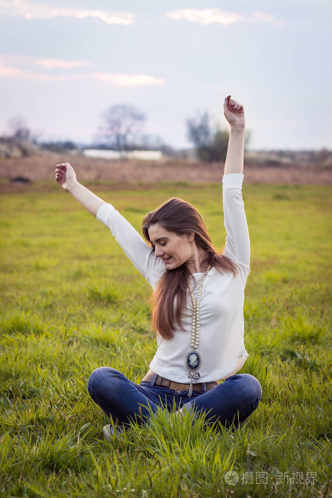 美丽的年轻女孩坐在草地上闭上眼睛双手举过头顶