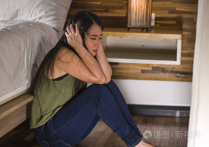 年轻忧郁悲伤的亚裔韩国女子坐在床旁卧室的地板上患有抑郁症和焦虑症