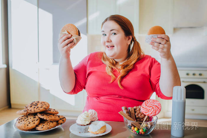 胖女人坐在厨房里吃垃圾食品手拿汉堡对着镜头微笑身体呈阳性大尺寸