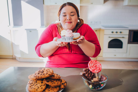 胖女人坐在厨房里吃着甜食.大码模特手里拿着小蛋糕看着.