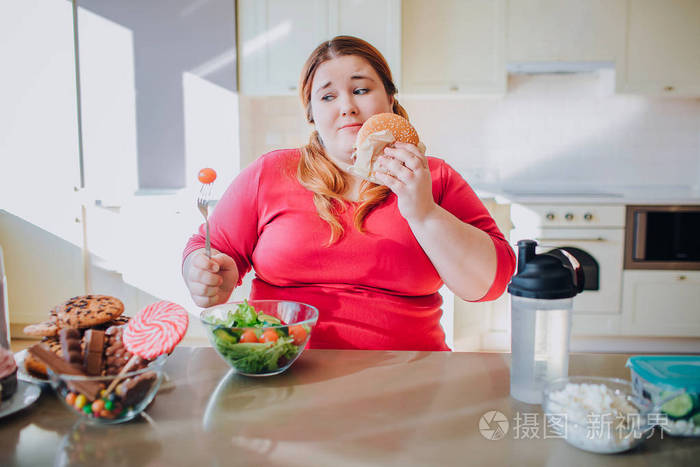 胖女人坐在厨房里吃垃圾食品.大码模特有不健康的饮食习惯.
