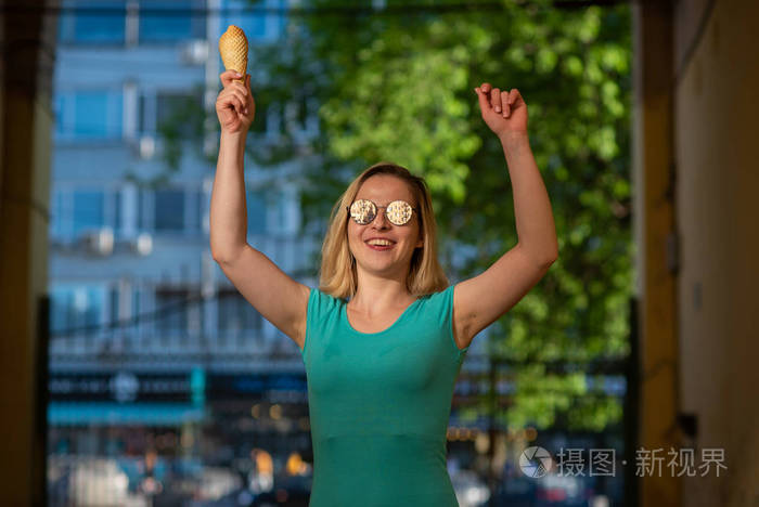 在一个温暖的夏天，一个穿着绿松石裙子的快乐女人站在院子里吃华夫饼。戴墨镜的金发美女边走边吃冰淇淋。夏季甜点。